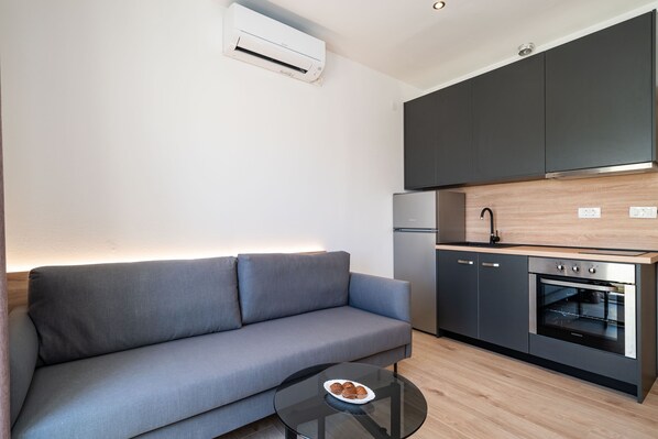 A modern kitchen with dark cabinets, a built-in oven, and a grey sofa. - Bacan Serviced Apartments (Konavle)