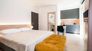Un dormitorio moderno con una cama grande, un escritorio pequeño y una kitchenette al fondo.