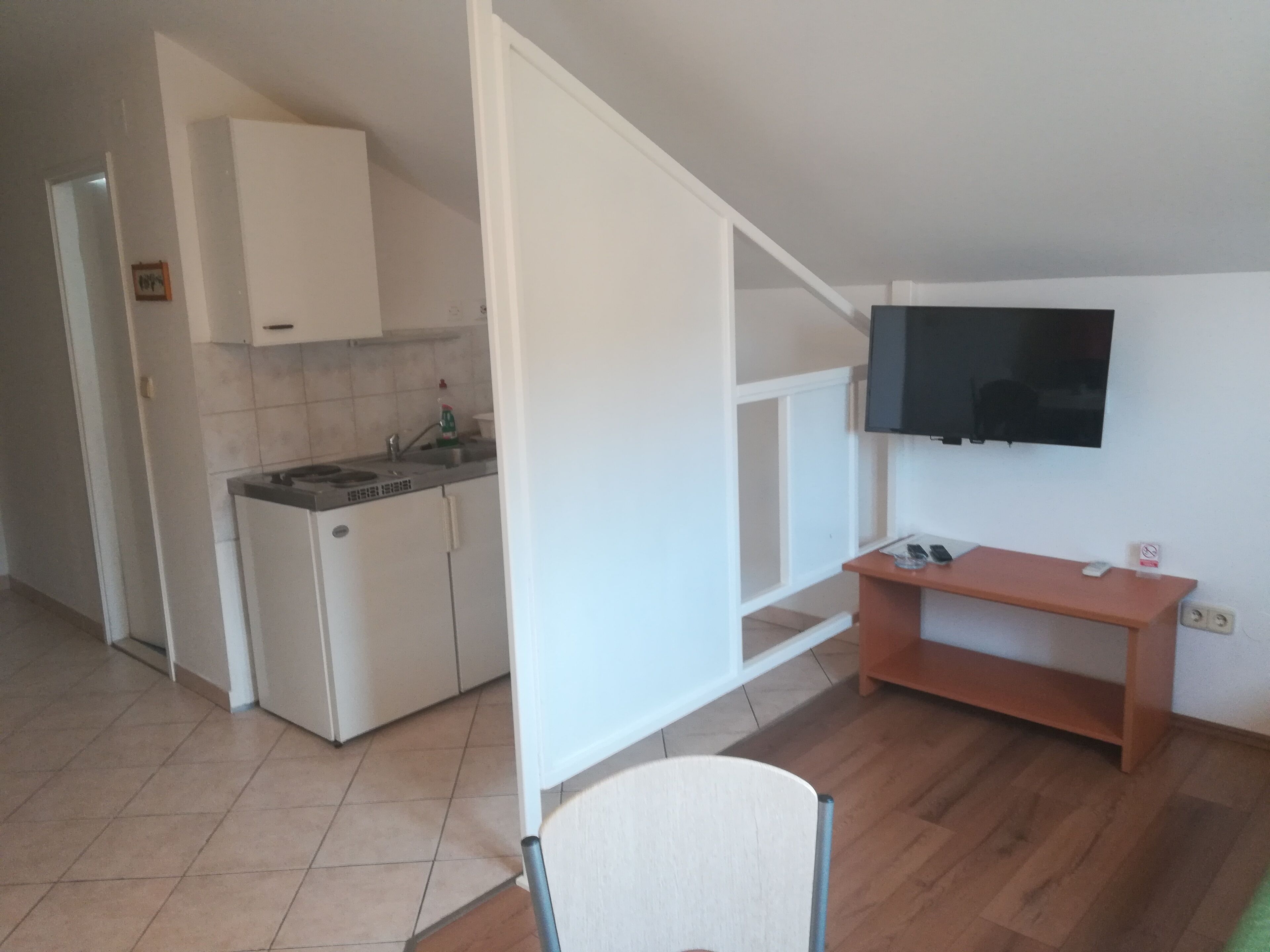 Cozinha compacta com armários brancos, fogão e pia. Uma televisão está instalada na parede acima de uma prateleira de madeira.