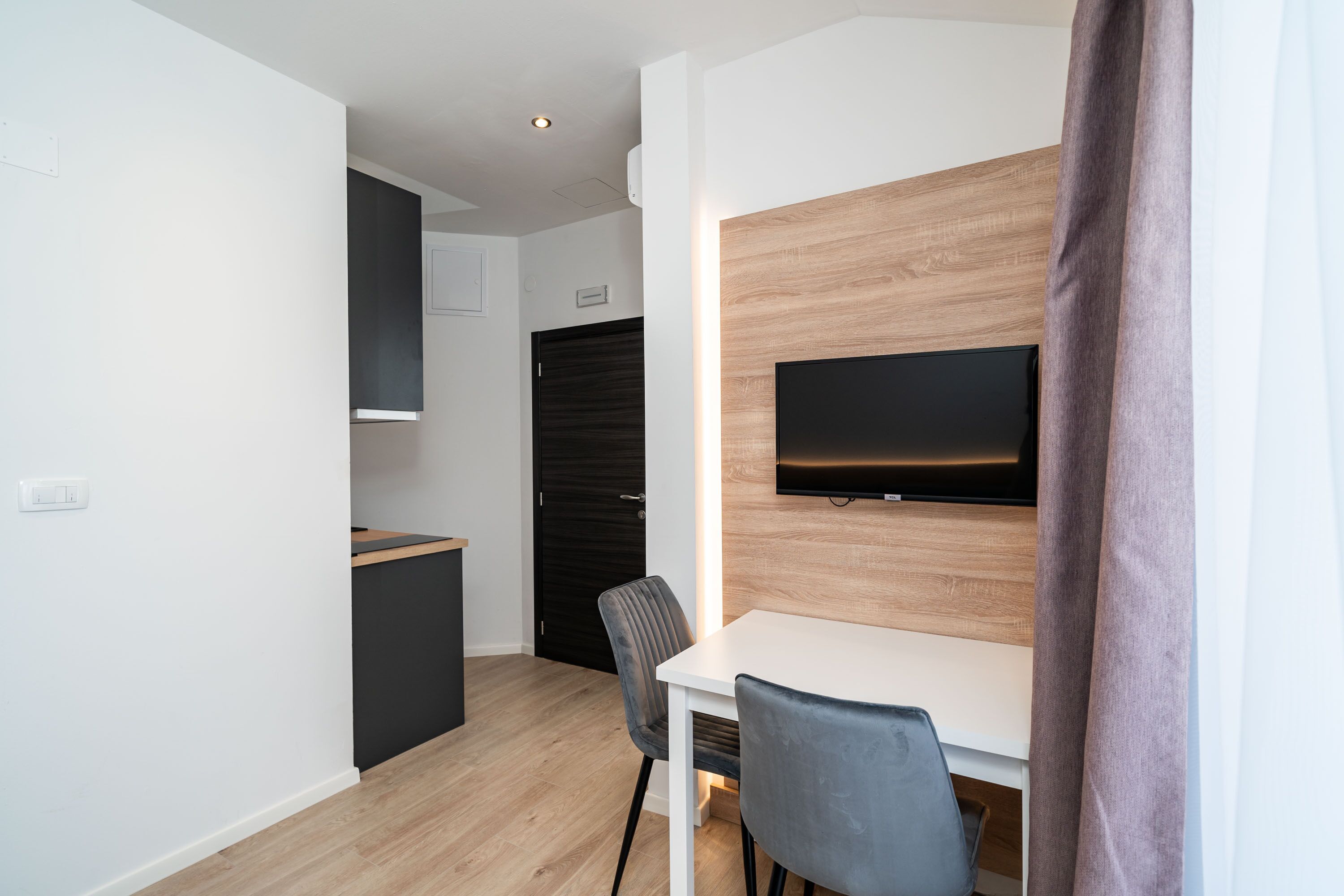 A modern, compact living space with a kitchenette, a dining area with a table and chairs, and a mounted TV on a wooden wall.