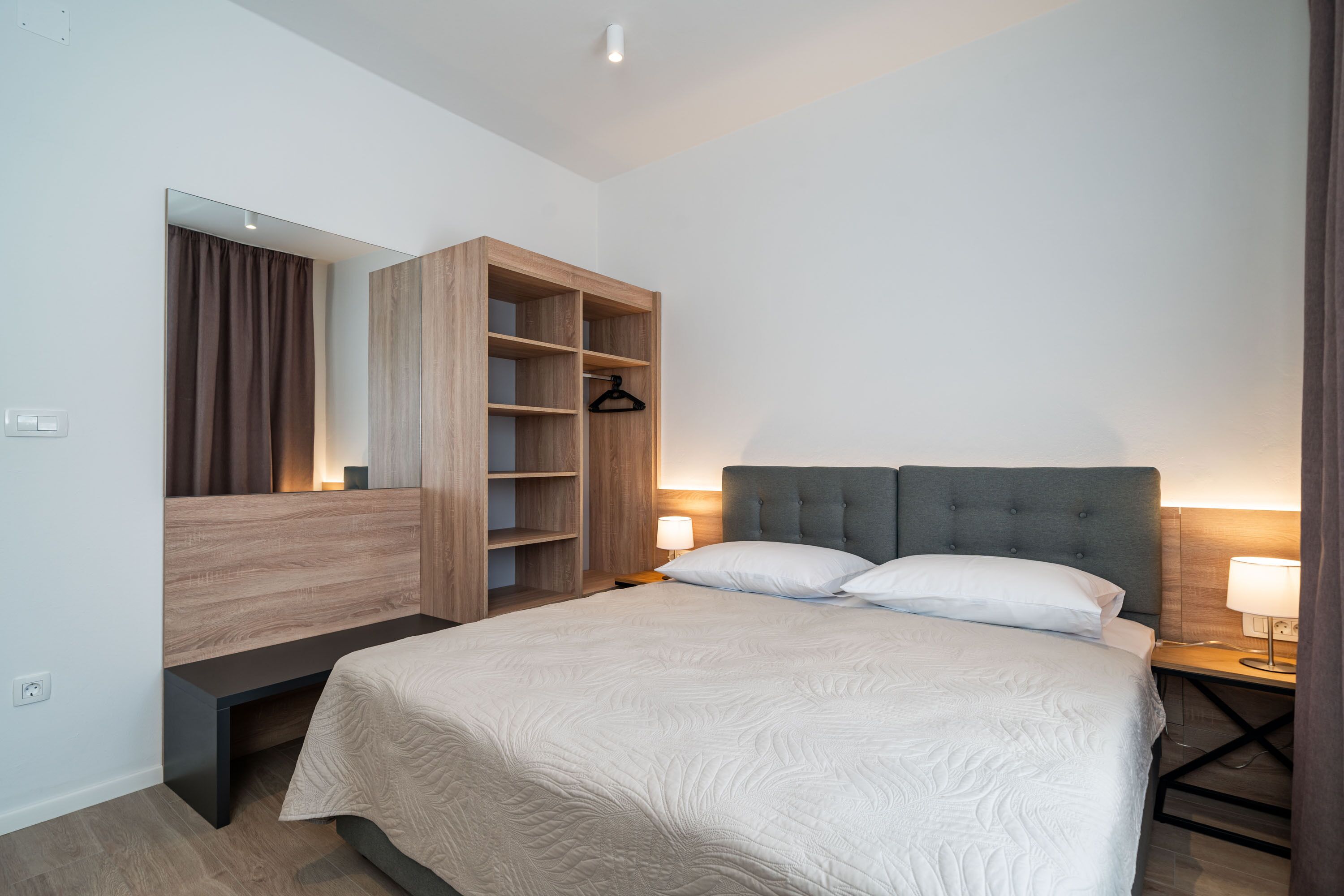 A modern bedroom with a large bed, a wooden wardrobe, a bench, and a bedside table with a lamp.