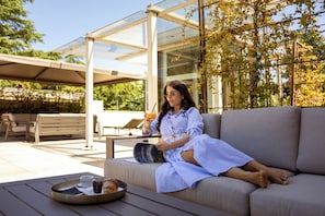 A woman sitting on a sofa with a tray of food and drink, reading a magazine, in a modern outdoor setting with a glass structure and greenery.