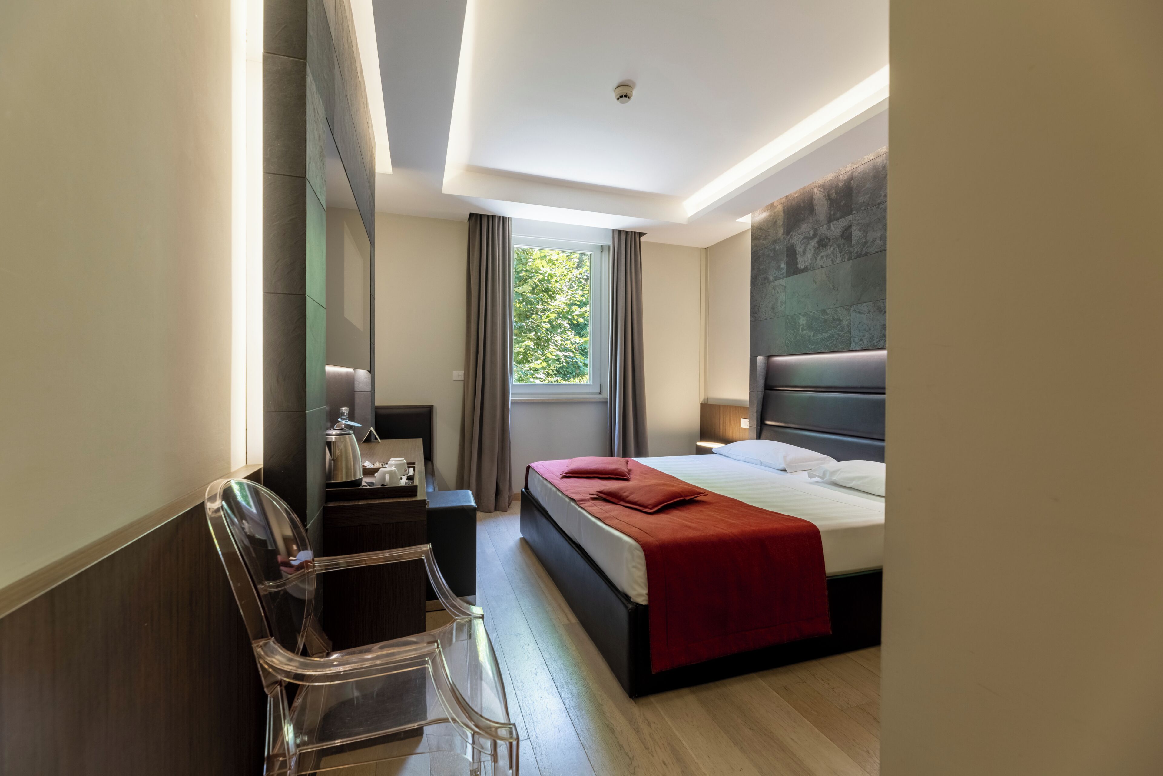 A modern hotel room with a bed, a desk, a chair, a window with curtains, and a view of greenery outside.