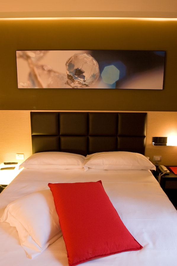 A hotel room with a bed, a red pillow, a headboard, and a wall-mounted artwork.