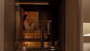 A modern sauna with glass walls, wooden benches, and a person sitting inside. - UNAHOTELS Villa dei Platani Foligno (Foligno)