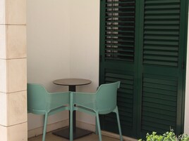 Uma pequena área externa com assentos, incluindo duas cadeiras turquesa e uma mesa redonda, localizada ao lado de uma porta verde com ripas horizontais.