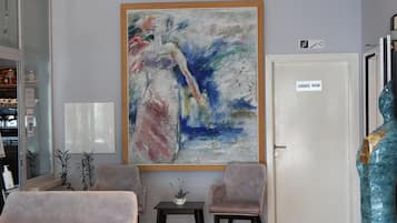 A room with a large abstract painting, a luggage room sign, two chairs, a small table with a plant, and a large sculpture.