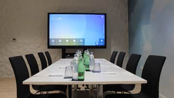Un salón de conferencias con una larga mesa, sillas negras, un televisor de pantalla plana en la pared y botellas de agua sobre la mesa.