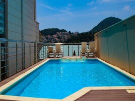 Piscina no terraço com água azul cristalina, rodeada por um deck de madeira e espreguiçadeiras bege.