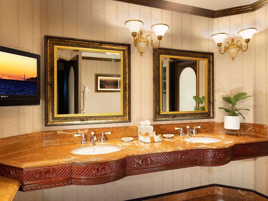 Une salle de bain avec deux lavabos, un grand miroir, des appliques murales et un téléviseur diffusant une image de coucher de soleil.