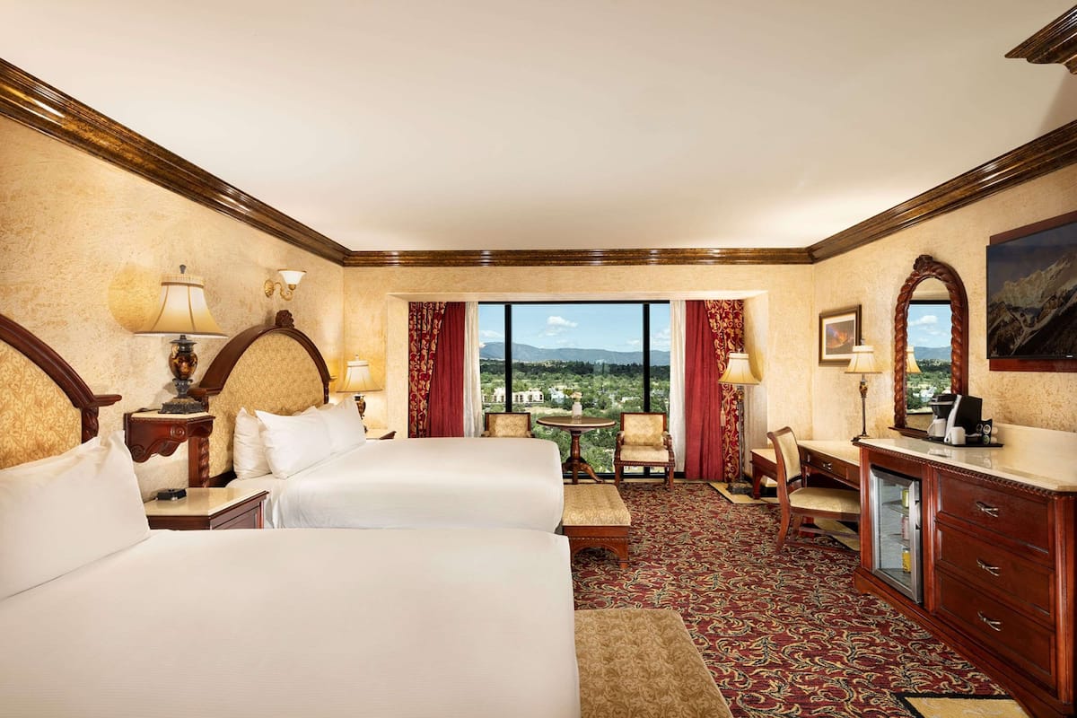 A hotel room with two beds, a desk, a chair, a small table, and a view of the city.