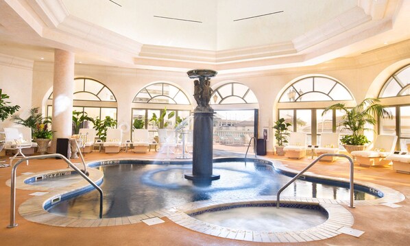 A large indoor pool with a central fountain, surrounded by lounge chairs and potted plants.