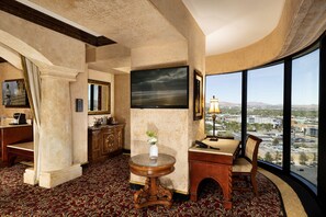 A hotel room with a large window, a round wooden table, a chair, a desk, a mirror, and a framed picture on the wall.
