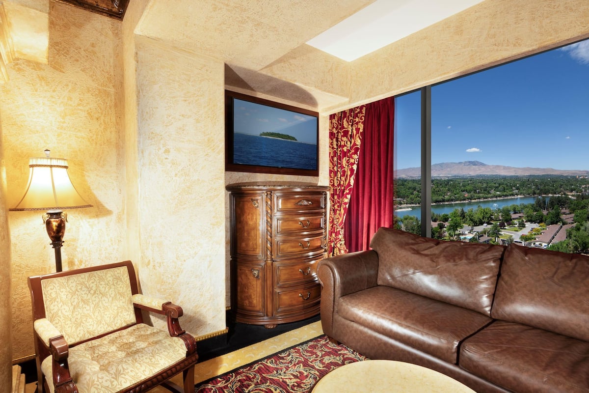 A room with a leather sofa, a wooden cabinet, a chair, a lamp, and a large window with a view of a landscape.