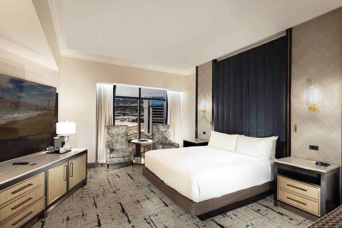 A hotel room with a bed, bedside tables, a desk with a lamp, a chair, a large window with a view, and a TV on the wall.