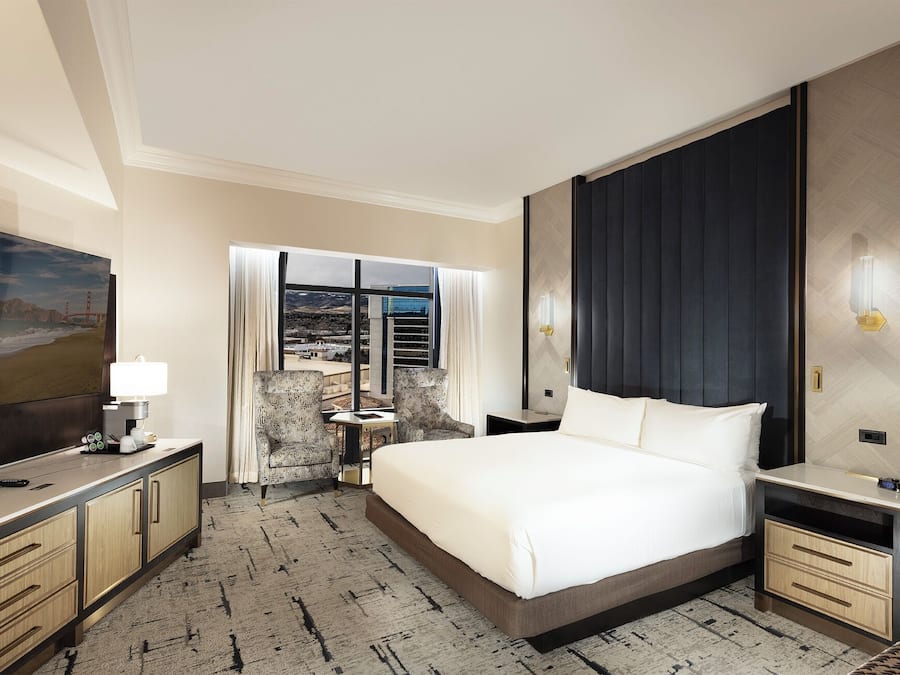 A hotel room with a bed, bedside tables, a desk with a lamp, a chair, a large window with a view, and a TV on the wall.