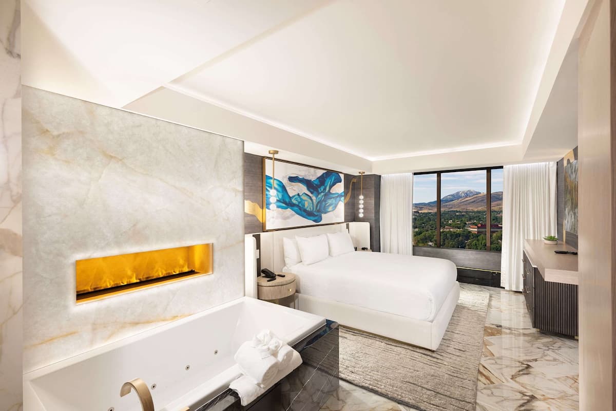 A modern hotel room with a large bed, a bathtub, a painting, and a view of the mountains.
