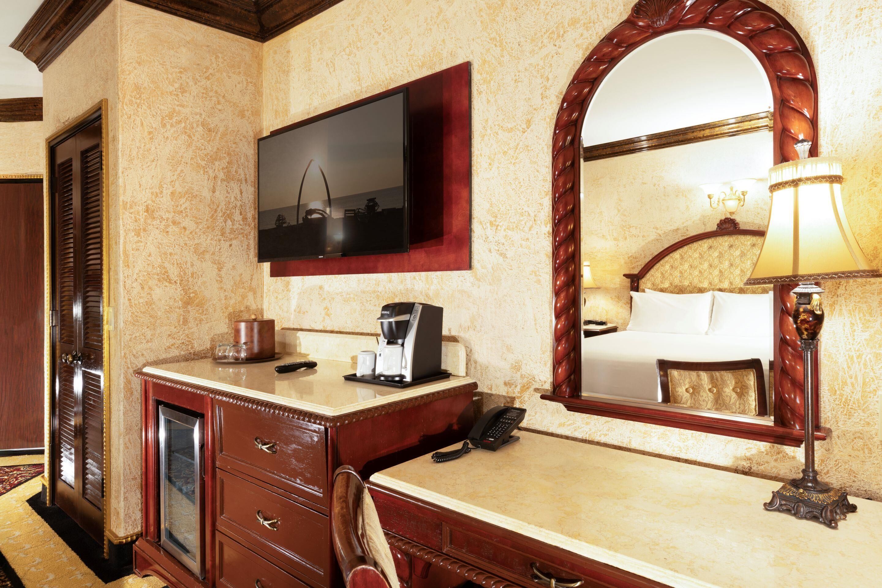 Ein Hotelzimmer mit einem Holztisch, einem darüber montierten Fernseher, einem Spiegel, der das Bett reflektiert, und einer Lampe auf dem Nachttisch.