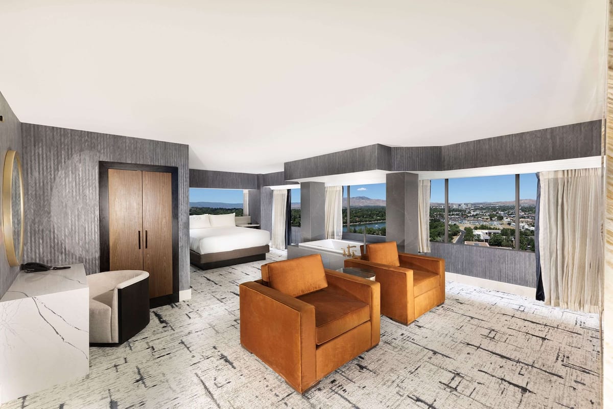 A modern hotel room with a large bed, a desk, two armchairs, and a view of a cityscape through large windows.