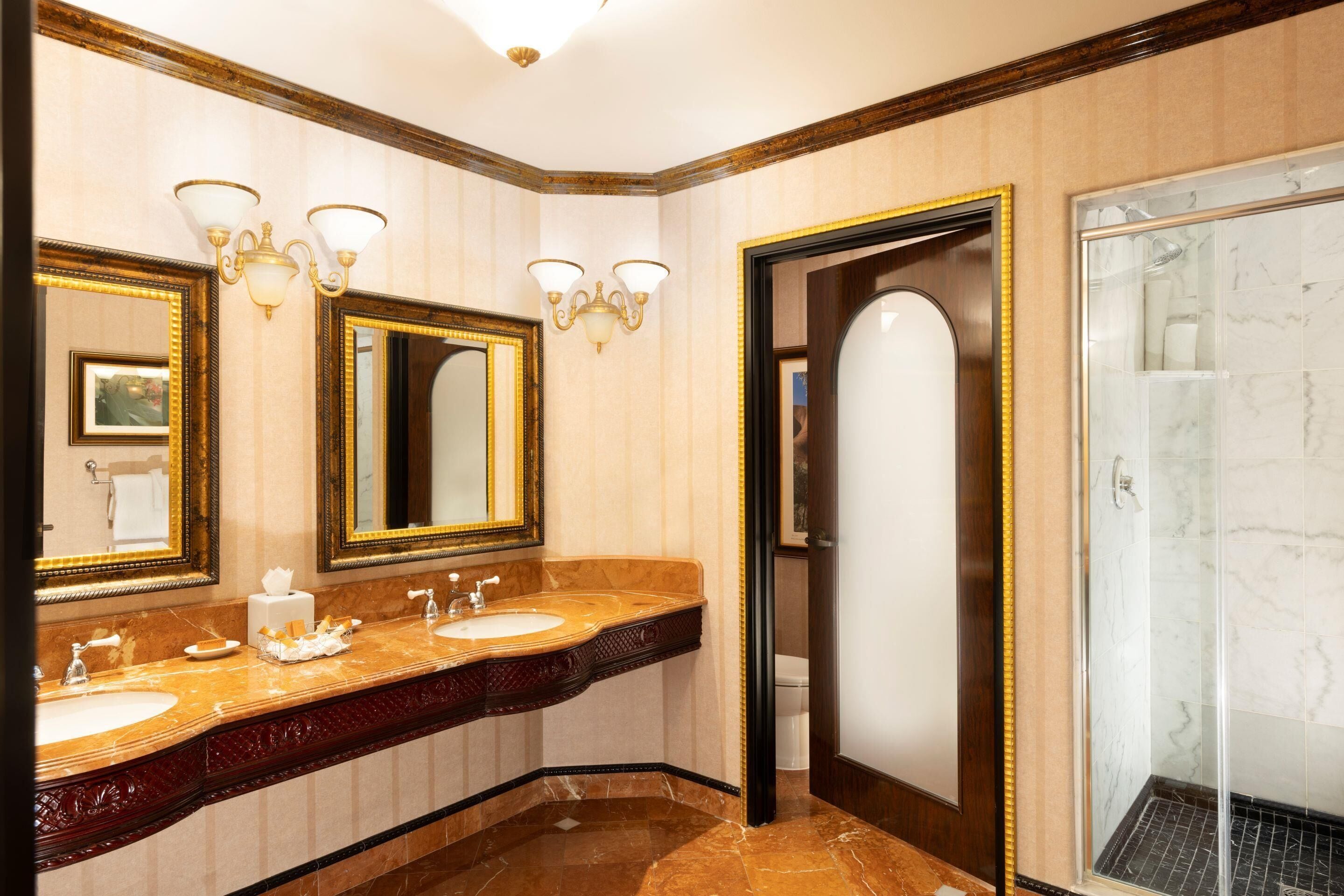 A hotel bathroom with two sinks, a large mirror, wall sconces, and a glass shower enclosure.