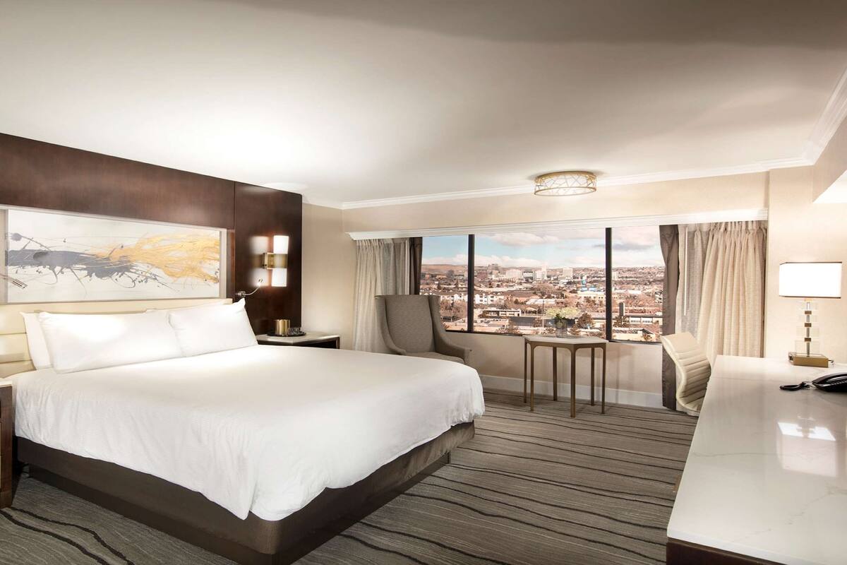 A hotel room with a large bed, a desk, a chair, a view of the city, and a painting on the wall.
