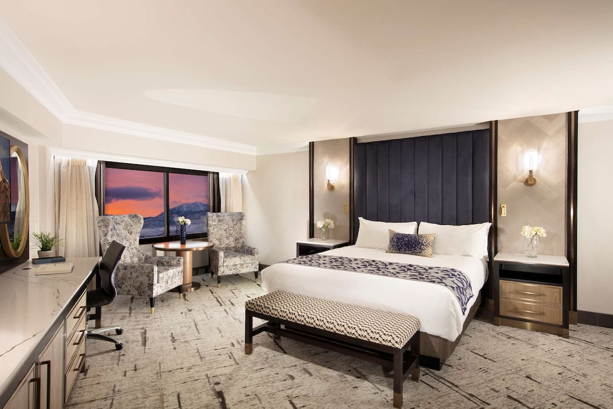 A hotel room with a large bed, a desk with a chair, a bench, a nightstand, and a view of a sunset through the window.
