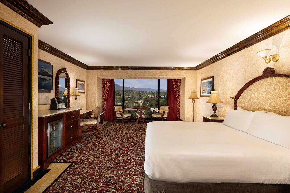 A hotel room with a large bed, a desk with a lamp, a chair, a small table, and a view of the mountains.
