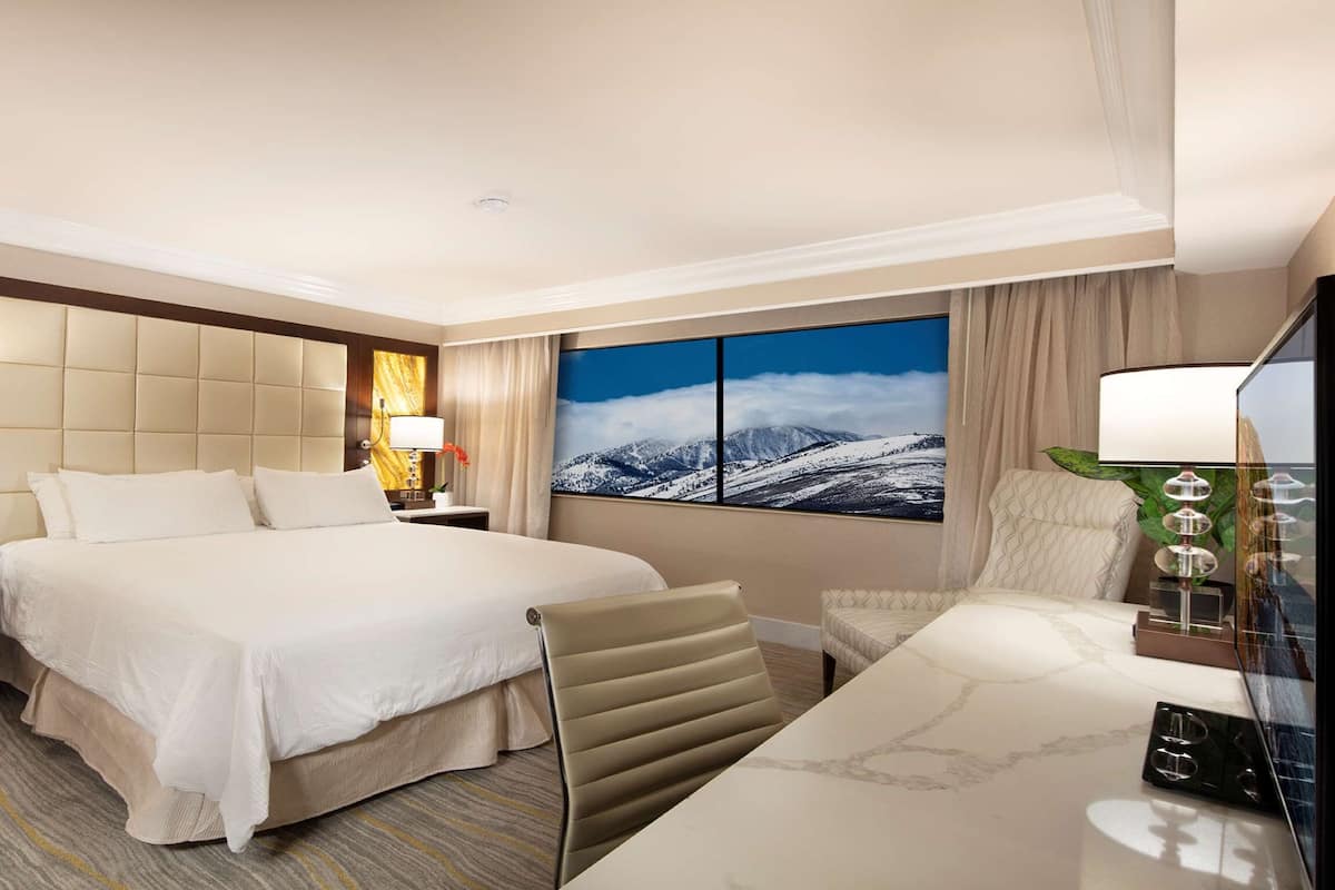 A hotel room with a large bed, a desk, a chair, a nightstand, a lamp, and a painting of a snowy mountain landscape.