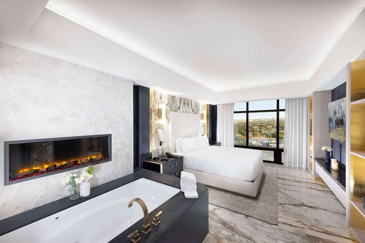 A modern hotel room with a large bed, a built-in fireplace, a large window with a city view, and a marble floor.