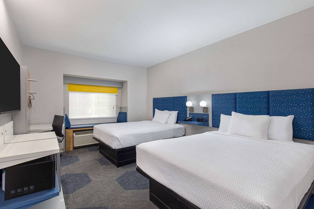 A hotel room with two beds, a desk, a chair, a TV, and a window with a yellow blind.