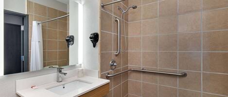 Une salle de bain avec un lavabo blanc, un miroir, une douche équipée d’une barre d’appui et un porte-serviettes.