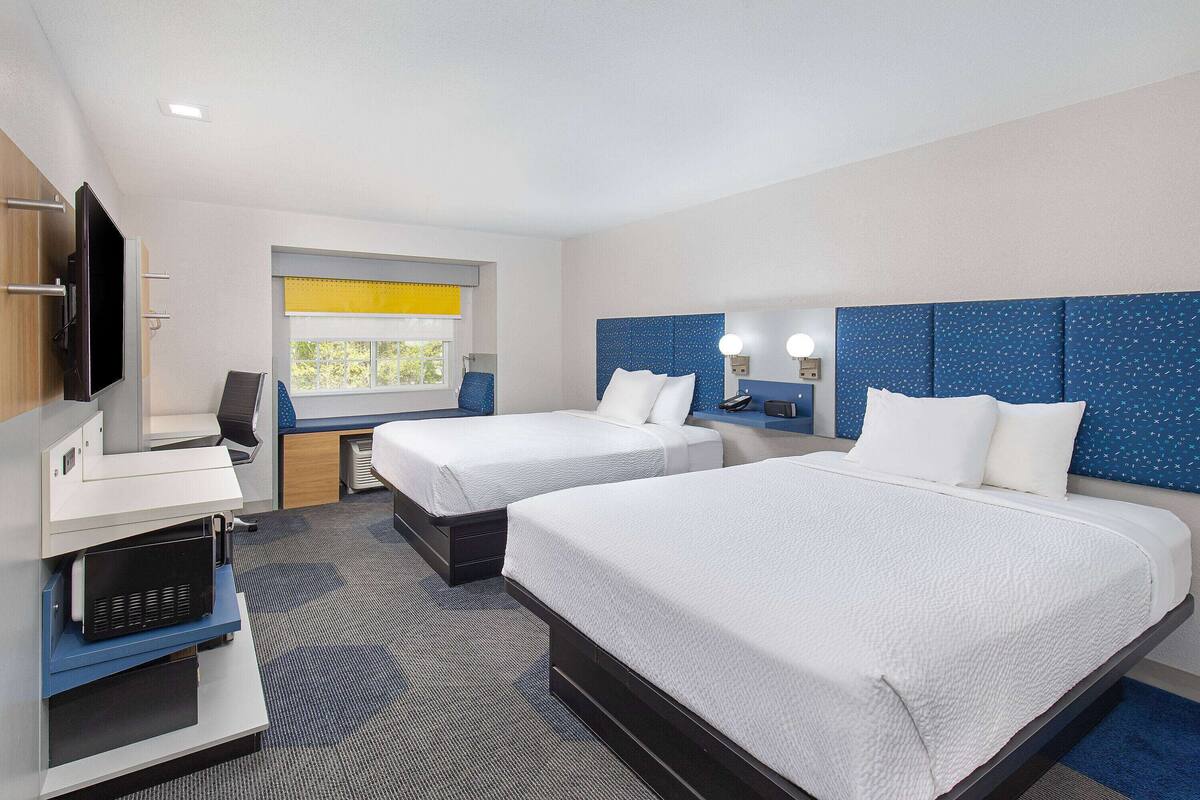 A hotel room with two beds, a desk, a television, and a window with yellow blinds.