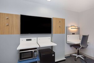 Una habitación de hotel moderna con televisión de pantalla plana, microondas, refrigerador pequeño, escritorio con silla y una estante pequeña.