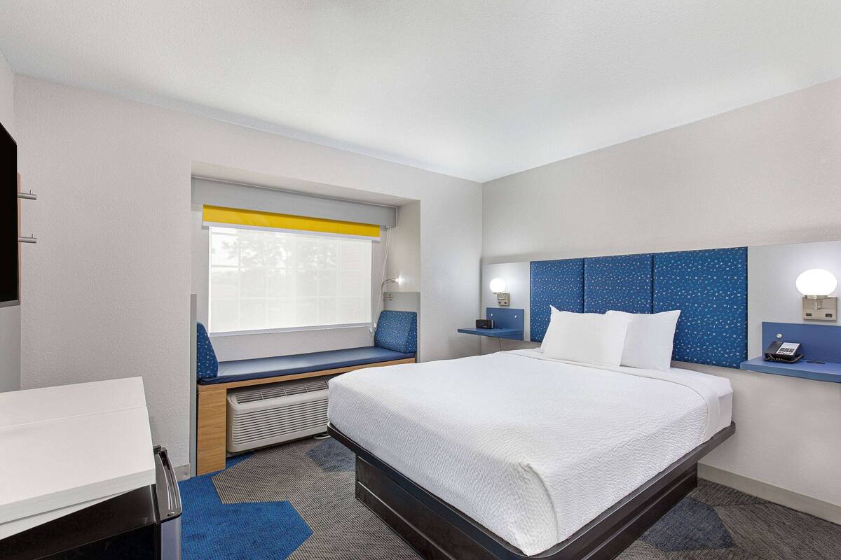 A hotel room with a bed, a desk, a window with a view, a blue headboard, and a bedside table with a lamp.