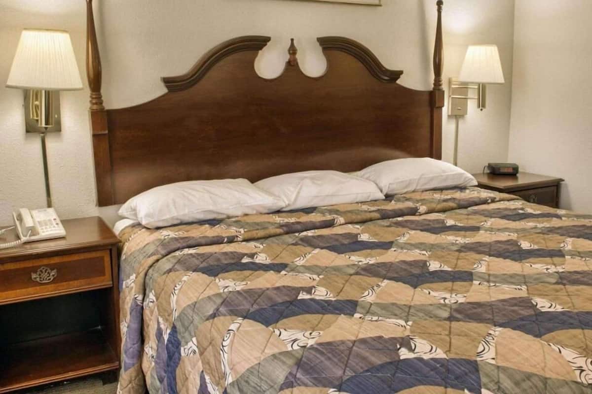 A hotel room with a wooden headboard, a quilted bedspread, two bedside lamps, a nightstand with a phone, and a painting on the wall.