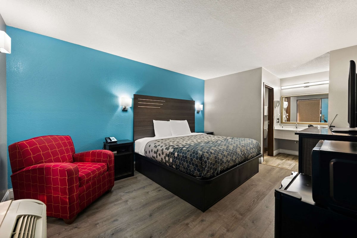 A hotel room with a large bed, a red armchair, a desk with a computer, and a bathroom visible through an open door.
