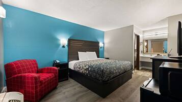A hotel room with a large bed, a red armchair, a desk with a computer, and a bathroom visible through an open door.