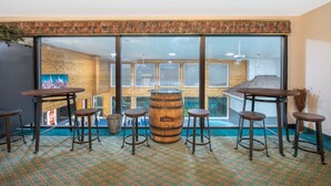 Un espace bar avec des tabourets en bois et un grand tonneau au centre, de grandes fenêtres et une vue sur une piscine.