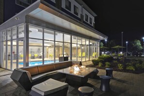 Un área de estar al aire libre con una chimenea, una piscina y un edificio acristalado con asientos en su interior.
