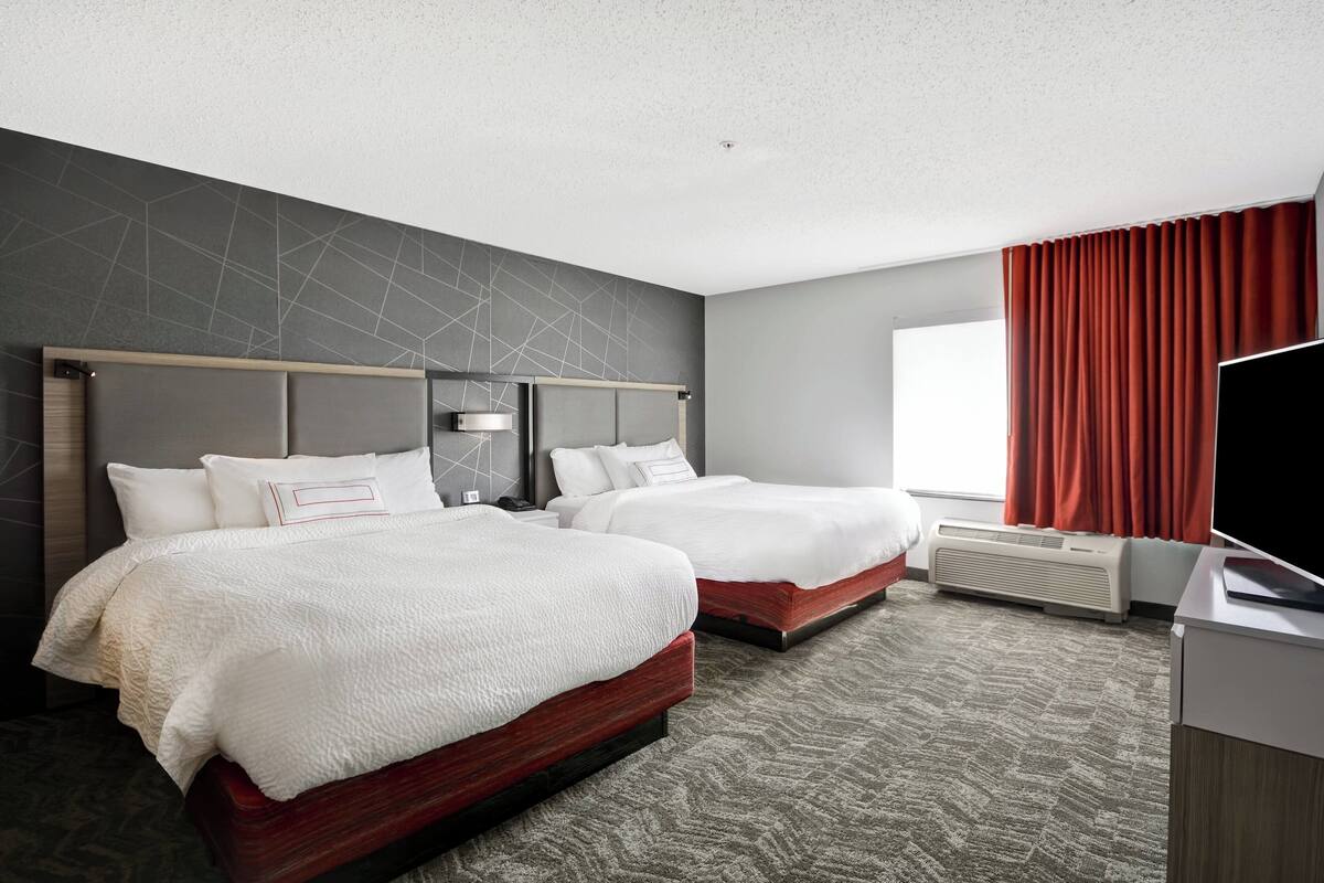 Ein Hotelzimmer mit zwei Betten, einem großen Fenster mit roten Vorhängen, einem Flachbildfernseher und einer Wand-Klimaanlage.