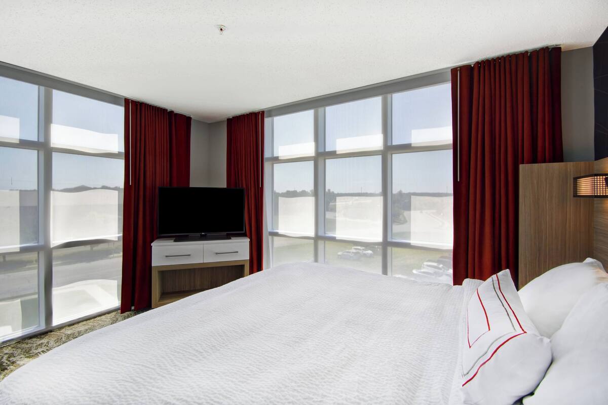 Ein Hotelzimmer mit einem großen Bett, einem Flachbildfernseher, einem Nachttisch aus Holz und Blick auf die Stadt durch große Fenster.