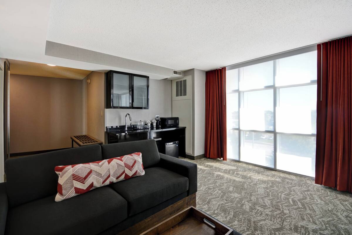 Ein kompaktes Hotelzimmer mit Küchenzeile, einem Sofa mit gemustertem Kissen und großen Fenstern mit roten Vorhängen.