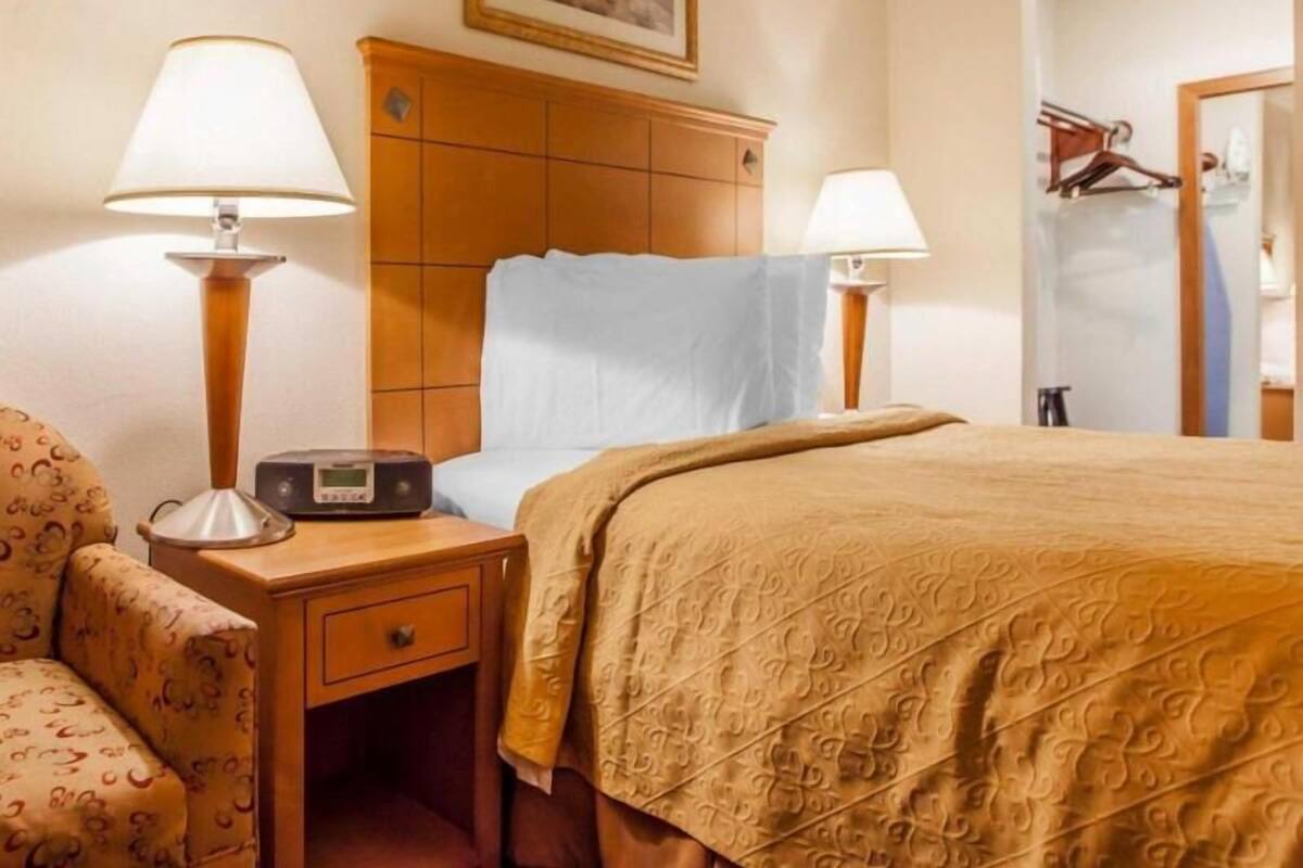 A hotel room with a bed, two bedside lamps, a nightstand with a radio, a chair, and a painting on the wall.
