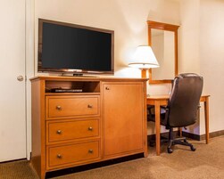 A hotel room with a wooden desk, a flat-screen TV, a lamp, a chair, and a mirror.