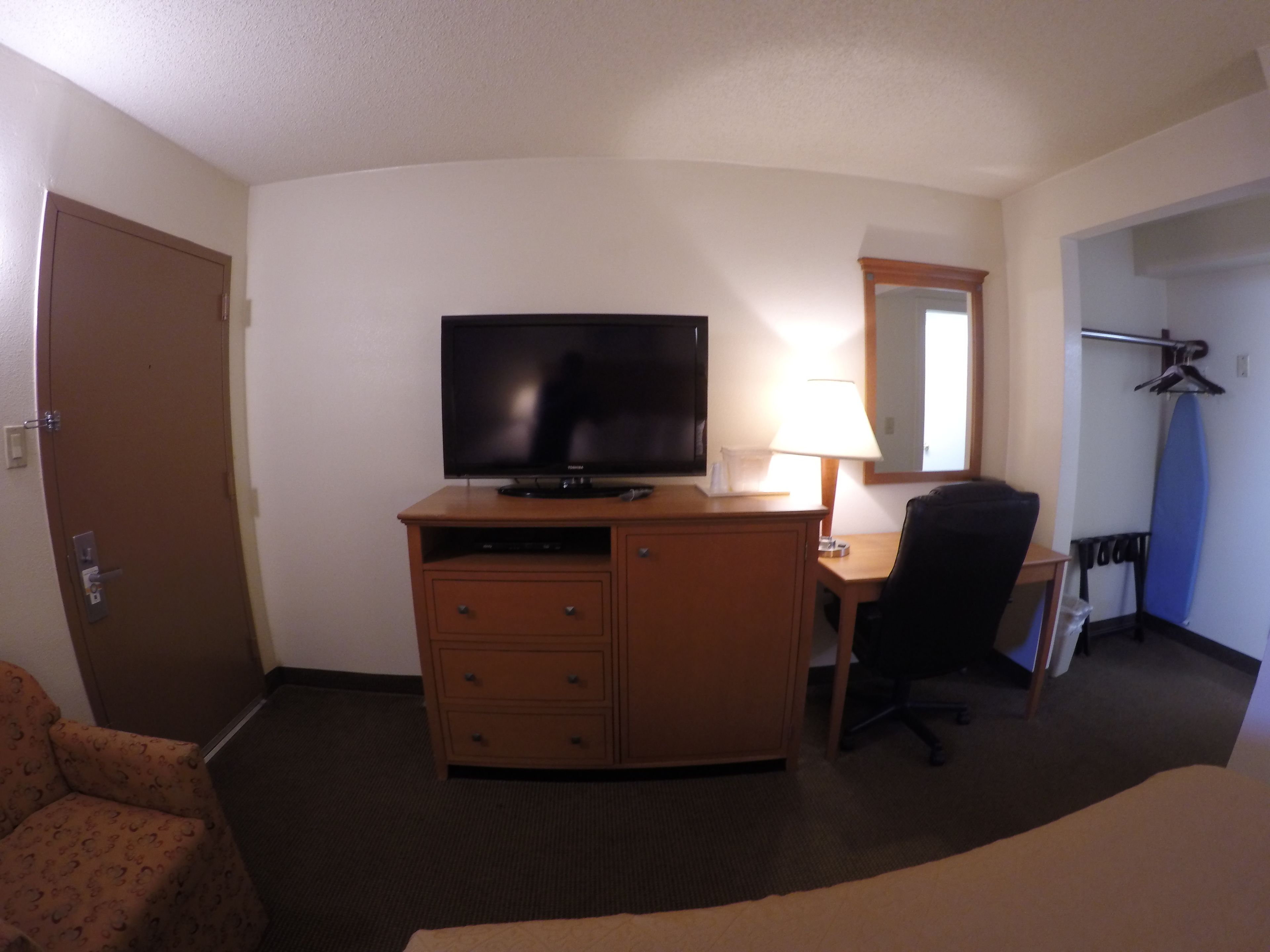 A hotel room with a TV on a wooden dresser, a desk with a lamp, a chair, and a bed.
