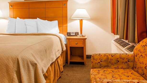 A hotel room with a bed, a nightstand with a lamp, a chair, and a painting on the wall. - Rodeway Inn Point Pleasant Beach (Point Pleasant Beach)