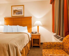 A hotel room with a bed, a nightstand with a lamp, a chair, and a painting on the wall. - Rodeway Inn Point Pleasant Beach (Point Pleasant Beach)