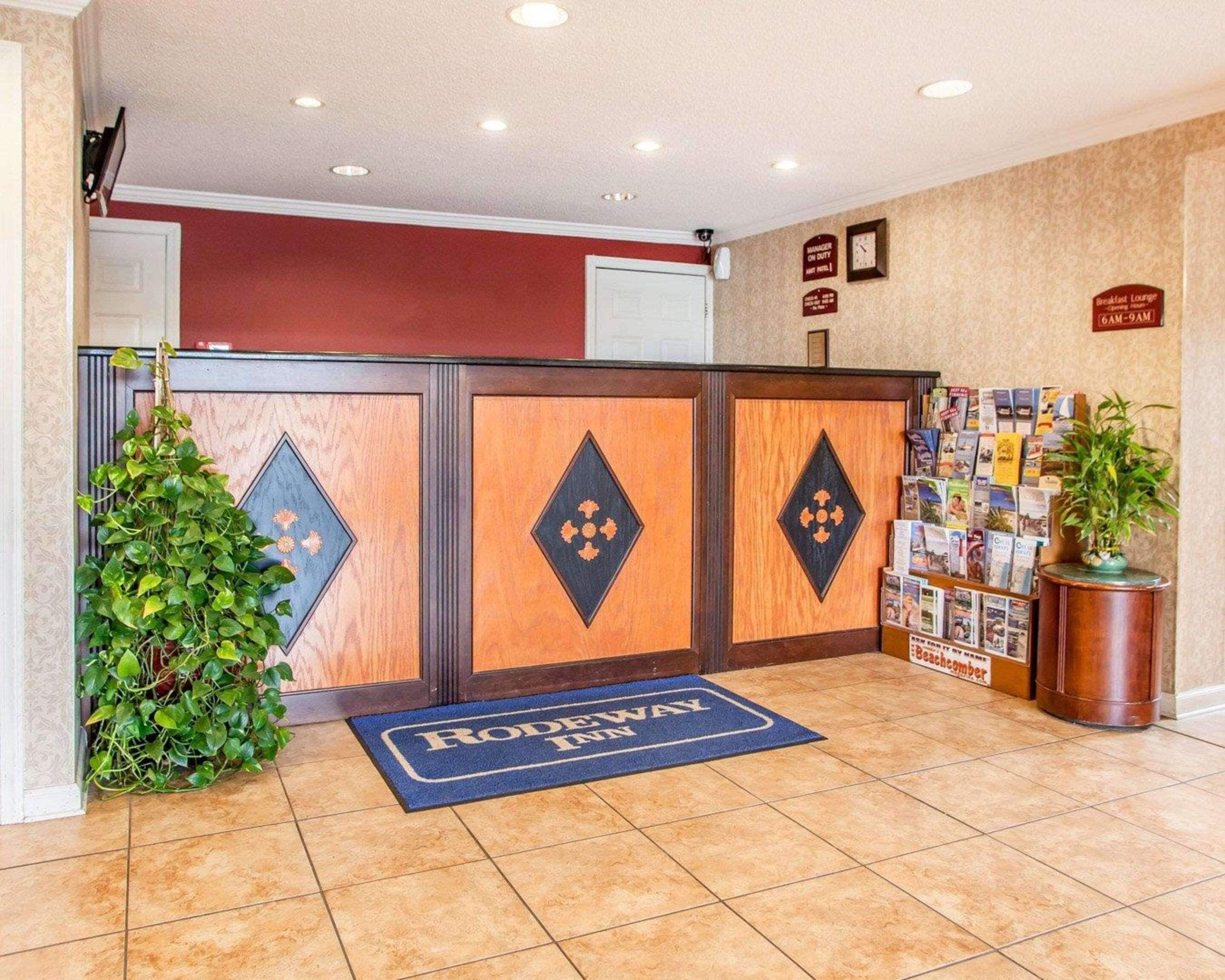 Un área de recepción con un mostrador de madera, un tapete azul con el texto “RODEWAY INN” y una pared con un tablón de anuncios y carteles.