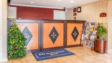 Un área de recepción con un mostrador de madera, un tapete azul con el texto “RODEWAY INN” y una pared con un tablón de anuncios y carteles.