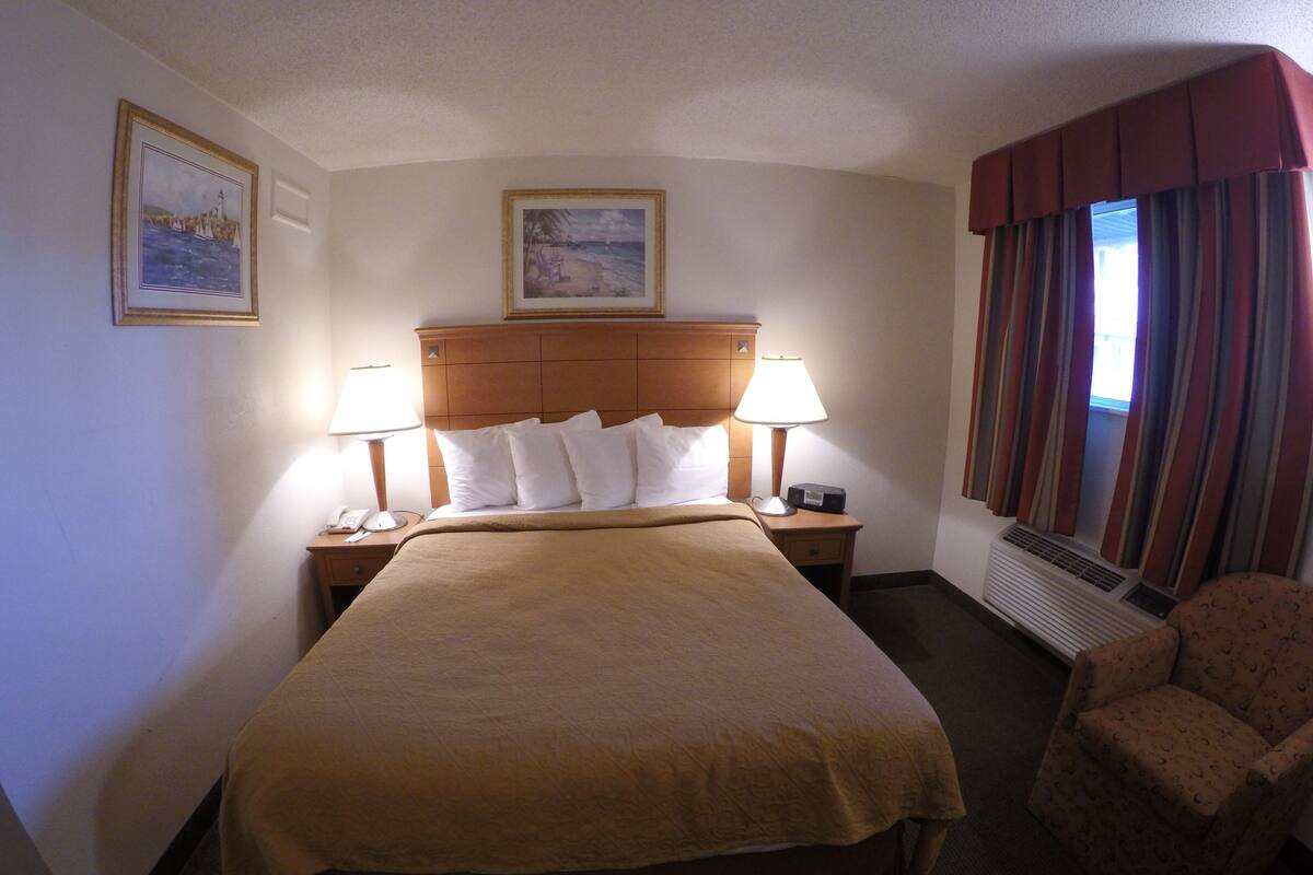 A hotel room with a large bed, two bedside lamps, a nightstand, a chair, a heater, and two framed pictures on the wall.